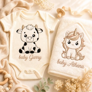 K&ouml;nnte beinhalten: Cremefarbener Baby-Strampler mit einer schwarz-wei&szlig;en Kuh-Grafik und dem Text "baby Garry". Ein passendes Kapuzenhandtuch zeigt ein Einhorn-Design und den Text "baby Athena". Ein Schnuller aus Holz und ein Bei&szlig;ring aus Holz sind ebenfalls im Bild.