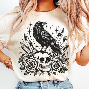 Pu&ograve; includere: T-shirt color crema con un design nero di un corvo appollaiato su un teschio circondato da rose e cristalli. Il design include stelle, creando un'estetica gotica e mistica. La grafica &egrave; centrata sulla maglietta.
