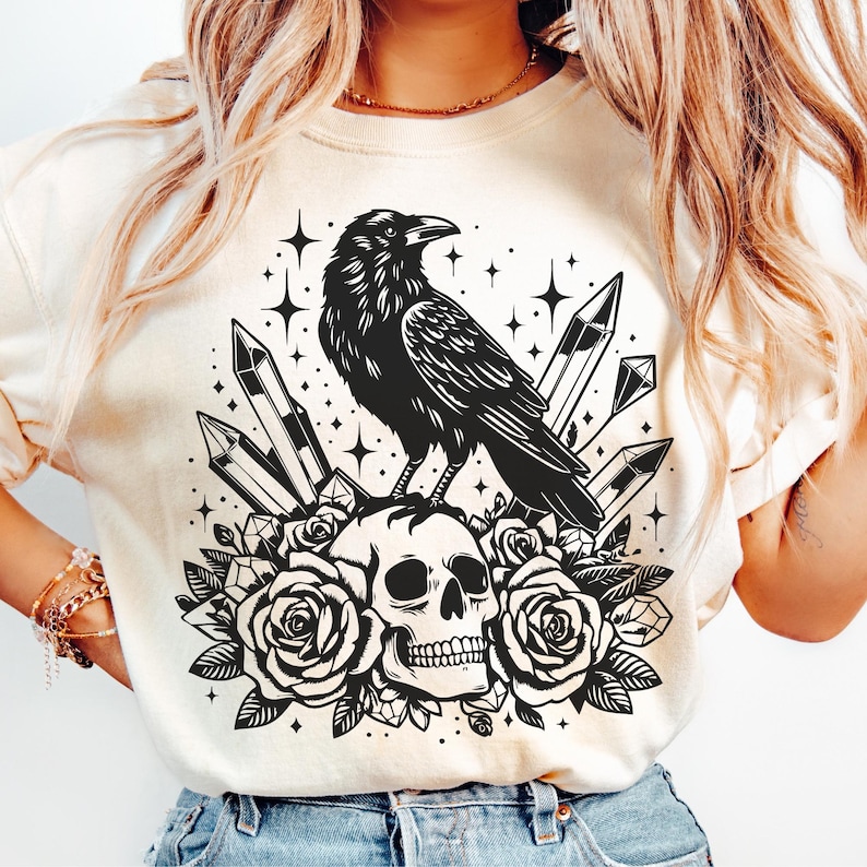 Pu&ograve; includere: T-shirt color crema con un design nero di un corvo appollaiato su un teschio circondato da rose e cristalli. Il design include stelle, creando un'estetica gotica e mistica. La grafica &egrave; centrata sulla maglietta.