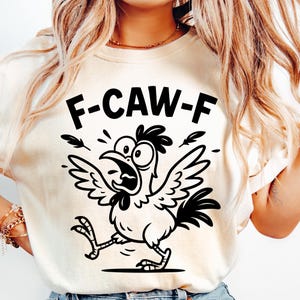 May include: Cream-colored t-shirt featuring a black and white cartoon chicken with the text "F-CAW-F". The chicken has a surprised expression, open beak, and outstretched wings. The design includes feather accents.