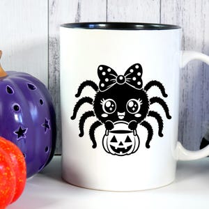 May include: A white ceramic mug with a black interior and handle, featuring a black graphic print of a cartoon spider with a polka-dot bow, large eyes, and a small smile, holding a jack-o'-lantern bucket for trick-or-treating.