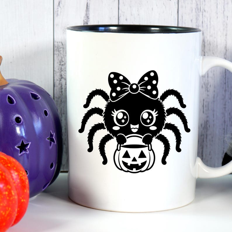 May include: A white ceramic mug with a black interior and handle, featuring a black graphic print of a cartoon spider with a polka-dot bow, large eyes, and a small smile, holding a jack-o'-lantern bucket for trick-or-treating.