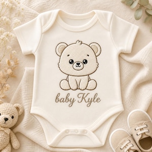 K&ouml;nnte beinhalten: Cremefarbener Baby-Body mit einem braunen, gestickten B&auml;ren-Design und dem Namen "baby Kyle". Der Body hat kurze &Auml;rmel und drei Druckkn&ouml;pfe am unteren Ende. Ein Paar beige Babyschuhe liegt neben dem Body.