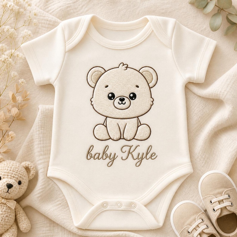 K&ouml;nnte beinhalten: Cremefarbener Baby-Body mit einem braunen, gestickten B&auml;ren-Design und dem Namen "baby Kyle". Der Body hat kurze &Auml;rmel und drei Druckkn&ouml;pfe am unteren Ende. Ein Paar beige Babyschuhe liegt neben dem Body.