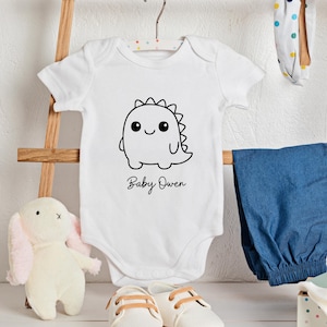 May include: White baby onesie with a black dinosaur graphic and the name "Baby Owen" in script. The onesie is hanging on a hanger with blue pants, a stuffed bunny, and baby shoes in the background.