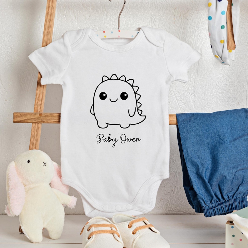 May include: White baby onesie with a black dinosaur graphic and the name "Baby Owen" in script. The onesie is hanging on a hanger with blue pants, a stuffed bunny, and baby shoes in the background.