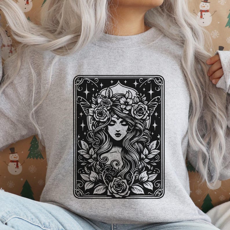 May include: A light gray sweatshirt featuring a black and white graphic of a woman with long hair, a floral crown, and wings. The design is framed with a decorative border and floral elements.
