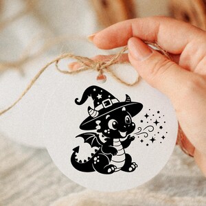 May include: A round white ornament featuring a black graphic illustration of a small dragon wearing a witch hat. The dragon is depicted with wings, a striped belly, and a tail, blowing out a stream of small stars. A brown twine string is attached.