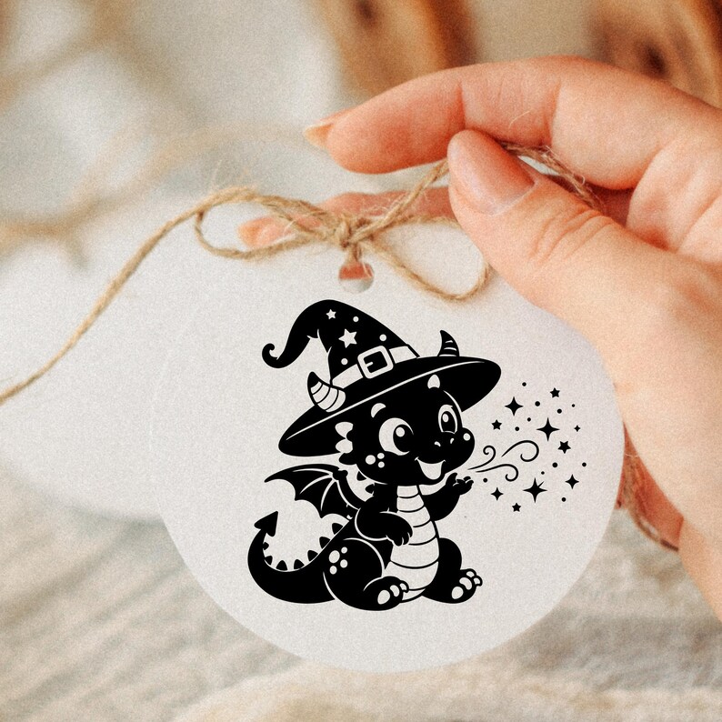 May include: A round white ornament featuring a black graphic illustration of a small dragon wearing a witch hat. The dragon is depicted with wings, a striped belly, and a tail, blowing out a stream of small stars. A brown twine string is attached.