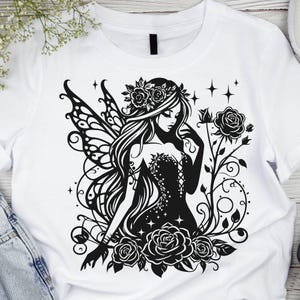 May include: White t-shirt featuring a black and white graphic of a fairy with floral accents. The fairy has long hair, a floral crown, and detailed wings. Roses and vines surround the fairy, creating a whimsical design.