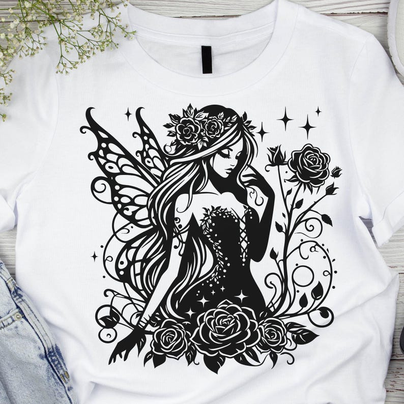May include: White t-shirt featuring a black and white graphic of a fairy with floral accents. The fairy has long hair, a floral crown, and detailed wings. Roses and vines surround the fairy, creating a whimsical design.