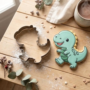 May include: A dinosaur-shaped cookie cutter next to a decorated dinosaur cookie. The cookie is light blue with a yellow spine, a red mouth, and black eyes. The cookie cutter is made of silver metal and is on a wooden surface.