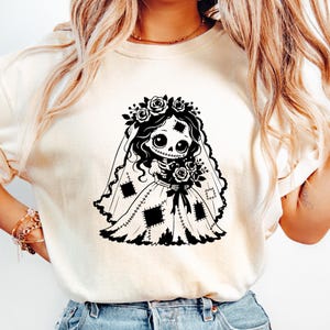 May include: A cream-colored t-shirt featuring a black graphic print of a skeletal bride character. The character wears a floral crown, a veil, and a dress with visible patch details, while holding a bouquet of roses in a stylized illustration.