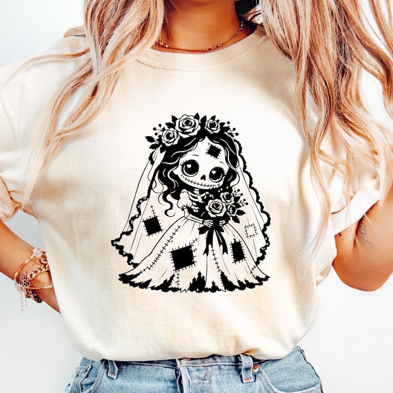 May include: A cream-colored t-shirt featuring a black graphic print of a skeletal bride character. The character wears a floral crown, a veil, and a dress with visible patch details, while holding a bouquet of roses in a stylized illustration.
