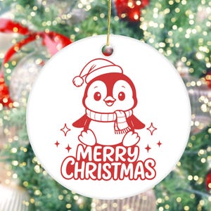 K&ouml;nnte beinhalten: Wei&szlig;er, runder Weihnachtsschmuck mit einem roten Pinguin, der einen Weihnachtsmannhut und einen Schal tr&auml;gt. Die Worte "MERRY CHRISTMAS" sind in roter Schrift unter dem Pinguin geschrieben. Der Schmuck hat eine goldene Schnur zum Aufh&auml;ngen.