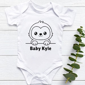May include: White baby onesie with a black and white cartoon sloth graphic and the text "Baby Kyle". The onesie is short-sleeved and has a simple, clean design, perfect for a newborn or infant.