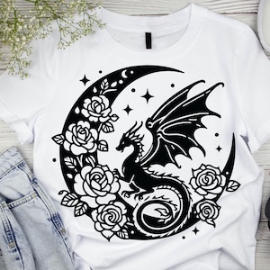 Puede incluir: Una camiseta blanca con un dise&ntilde;o negro de un drag&oacute;n enroscado alrededor de una luna creciente con estrellas y rosas.