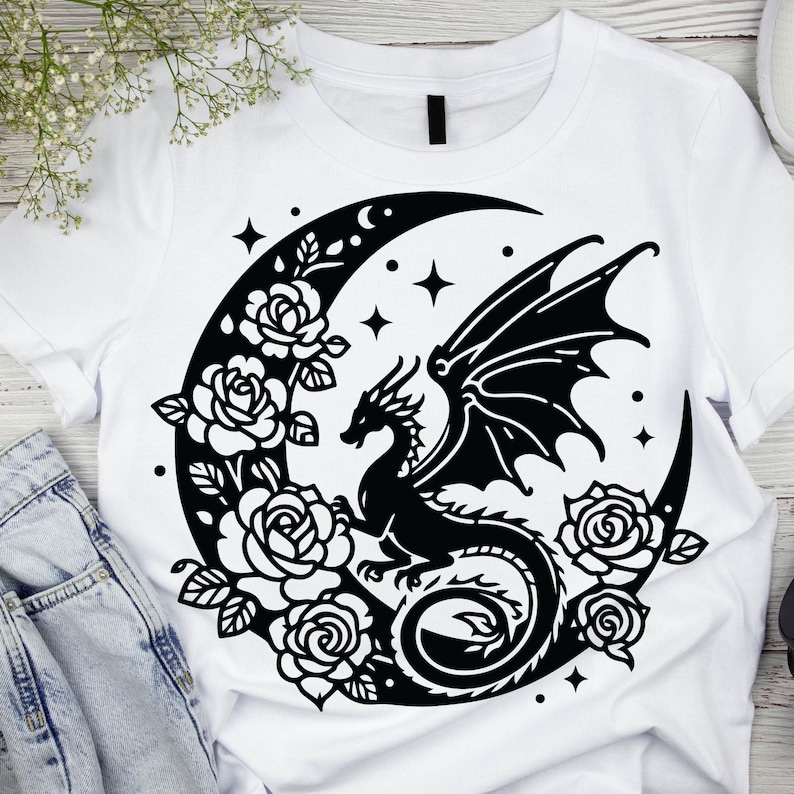 Puede incluir: Una camiseta blanca con un dise&ntilde;o negro de un drag&oacute;n enroscado alrededor de una luna creciente con estrellas y rosas.