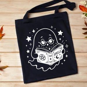 May include: A black tote bag featuring a white graphic of a ghost wearing round glasses and reading an open book. The book cover displays the word SPELLS, a pentagram, and a crescent moon. The design is surrounded by various sizes of white stars.