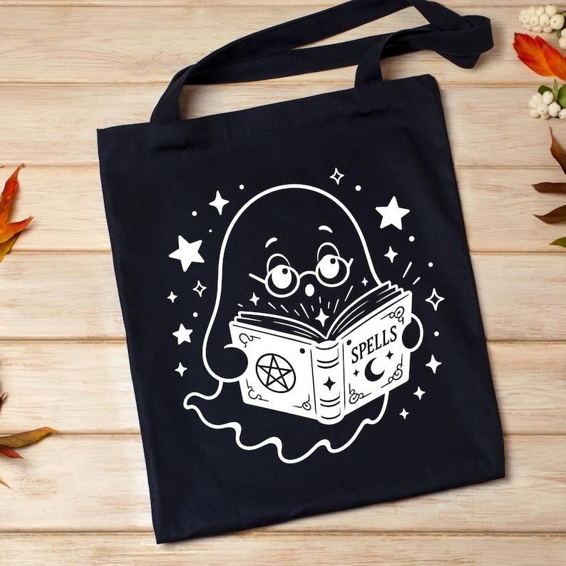 May include: A black tote bag featuring a white graphic of a ghost wearing round glasses and reading an open book. The book cover displays the word SPELLS, a pentagram, and a crescent moon. The design is surrounded by various sizes of white stars.