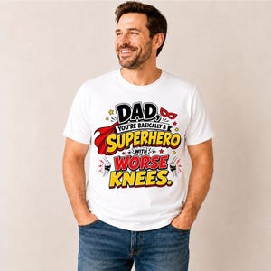 May include: A white t-shirt with a colorful graphic that reads "DAD, YOU'RE BASICALLY A SUPERHERO WITH WORSE KNEES." The design includes a red cape, a superhero mask, and yellow text. The person is wearing blue jeans.