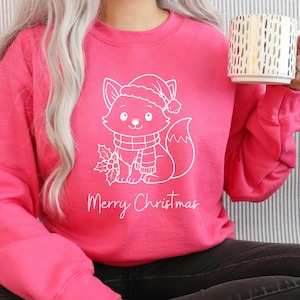 May include: A bright pink sweatshirt featuring a white cartoon fox wearing a Santa hat and scarf, with holly and the text "Merry Christmas." The sweatshirt is a festive holiday design.