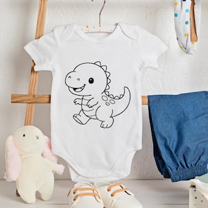 May include: White baby onesie with a black outline cartoon dinosaur design. The onesie is hanging on a wooden hanger. Other baby items include blue pants, white shoes, and a stuffed bunny.