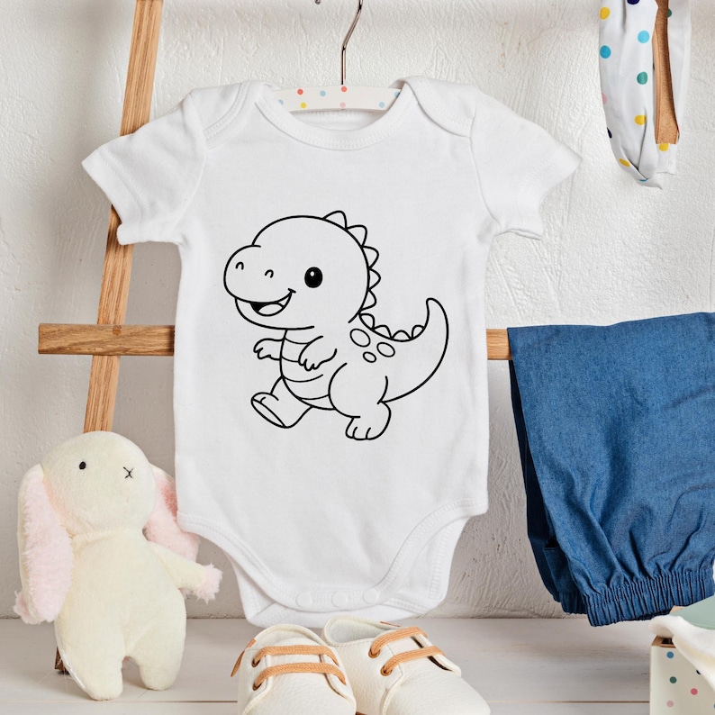 May include: White baby onesie with a black outline cartoon dinosaur design. The onesie is hanging on a wooden hanger. Other baby items include blue pants, white shoes, and a stuffed bunny.