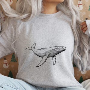 K&ouml;nnte beinhalten: Ein hellgraues Sweatshirt mit einer schwarz-wei&szlig;en Illustration eines Wals. Der Wal wird in einer Schwimmposition gezeigt, mit seinen Flossen und seinem Schwanz ausgestreckt. Das Sweatshirt ist aus weichem Material.