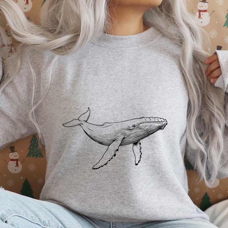 K&ouml;nnte beinhalten: Ein hellgraues Sweatshirt mit einer schwarz-wei&szlig;en Illustration eines Wals. Der Wal wird in einer Schwimmposition gezeigt, mit seinen Flossen und seinem Schwanz ausgestreckt. Das Sweatshirt ist aus weichem Material.