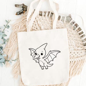 May include: Cream-colored tote bag featuring a black and white cartoon pterodactyl design. The pterodactyl has large wings and a friendly expression. The bag has two long handles and is made of a sturdy fabric, suitable for carrying various items.
