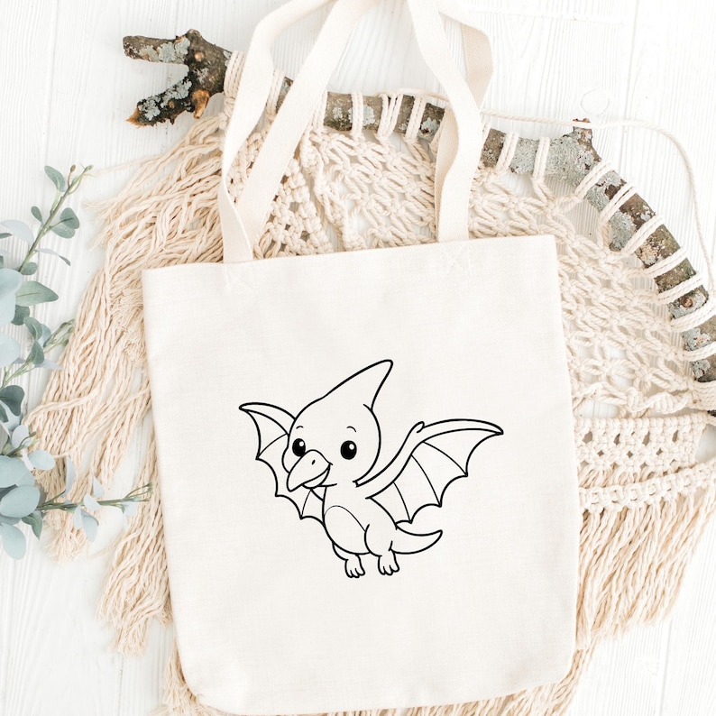 May include: Cream-colored tote bag featuring a black and white cartoon pterodactyl design. The pterodactyl has large wings and a friendly expression. The bag has two long handles and is made of a sturdy fabric, suitable for carrying various items.