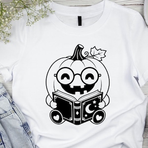 May include: A white t-shirt featuring a black graphic print of a smiling pumpkin character wearing round glasses. The pumpkin is holding an open book decorated with a crescent moon and stars. The design includes a single leaf on the pumpkin stem.