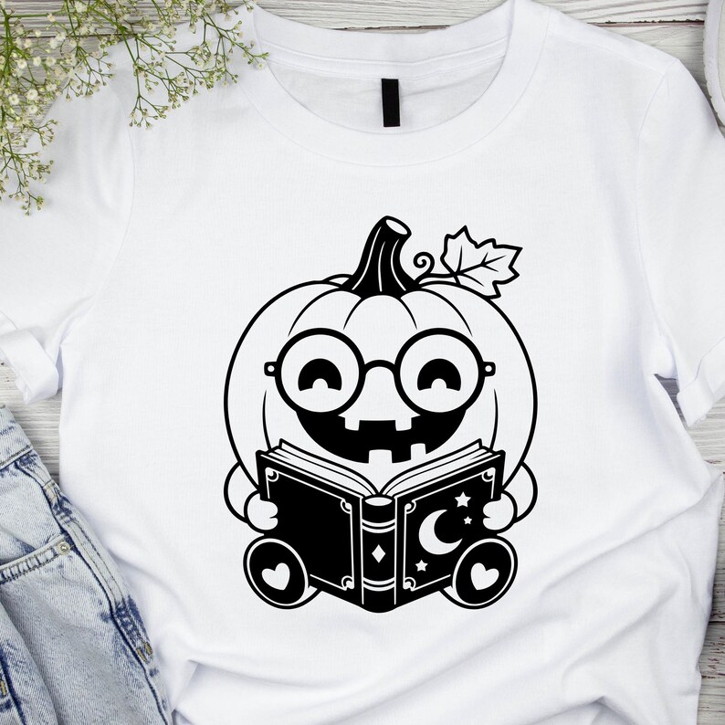 May include: A white t-shirt featuring a black graphic print of a smiling pumpkin character wearing round glasses. The pumpkin is holding an open book decorated with a crescent moon and stars. The design includes a single leaf on the pumpkin stem.