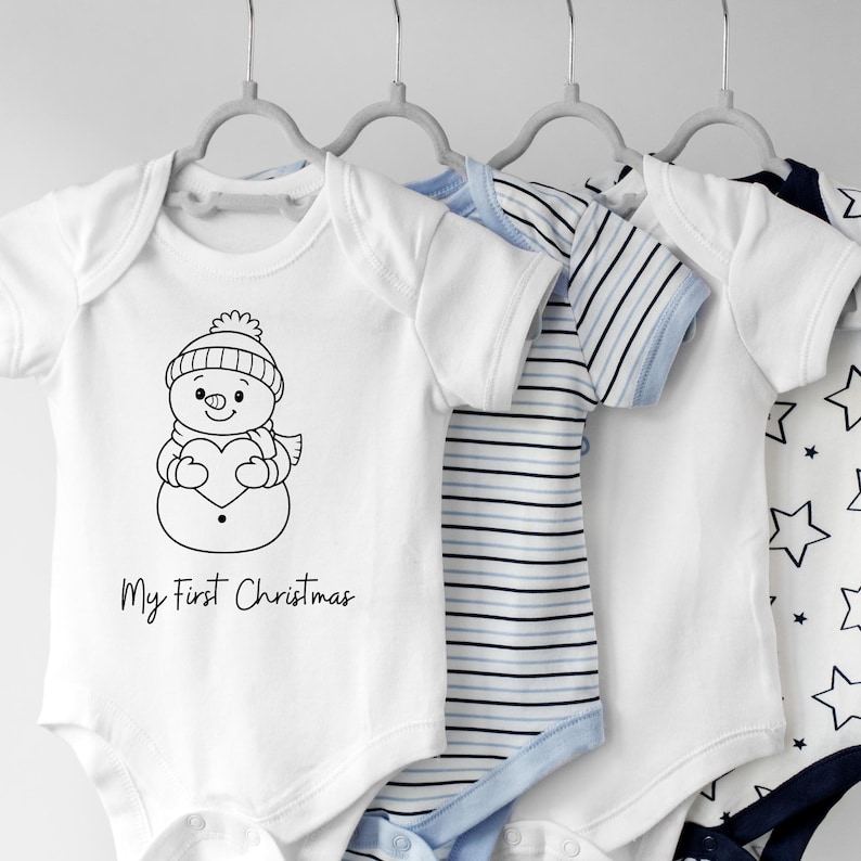May include: White baby onesies hanging on gray hangers. One onesie features a snowman graphic and the text "My First Christmas." Other onesies have blue and white striped and star patterns. The onesies are designed for infants.