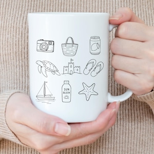 Pu&ograve; includere: Tazza in ceramica bianca con disegni a tratto nero a tema spiaggia: macchina fotografica, borsa da spiaggia, lattina, tartaruga, castello di sabbia, infradito, barca a vela, crema solare e stella marina. La tazza &egrave; tenuta da una persona.