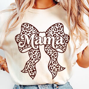 Op de afbeelding: Crèmekleurig T-shirt met een groot strikdesign met een luipaardprint. Het woord "Mama" staat in een sierlijk lettertype over het midden van de strik geschreven. Het shirt heeft een casual stijl.