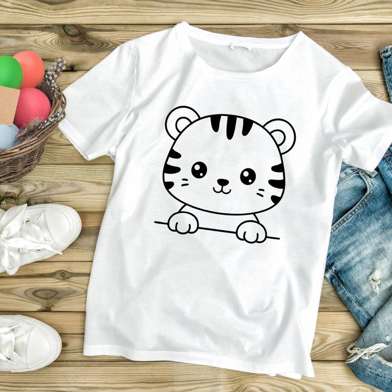 May include: White t-shirt featuring a black and white cartoon tiger design. The tiger has stripes, a cute face, and is peeking over a line. The shirt is displayed on a wooden surface with other items in the background.