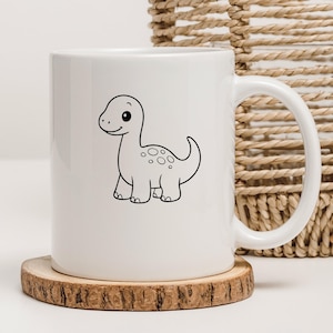May include: White ceramic mug featuring a black and white cartoon dinosaur illustration. The dinosaur has a friendly expression and is decorated with spots. The mug sits on a wooden coaster.