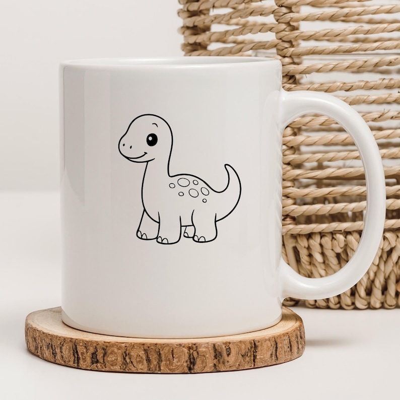 May include: White ceramic mug featuring a black and white cartoon dinosaur illustration. The dinosaur has a friendly expression and is decorated with spots. The mug sits on a wooden coaster.