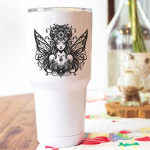May include: White insulated tumbler featuring a black illustration of a fairy with detailed wings, a floral crown, and ornate jewelry. The design is centered on the tumbler, which has a silver rim and a white body.