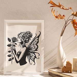 May include: A framed black and white illustration of a fairy smelling a rose. The fairy has large detailed wings and is adorned with roses in her hair. The artwork is in a white frame and is displayed on a white surface.
