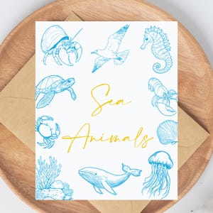 K&ouml;nnte beinhalten: Eine wei&szlig;e Karte mit blauen Illustrationen von Meerestieren, darunter eine Krabbe, eine Schildkr&ouml;te und ein Wal. Die Worte "Sea Animals" sind in gelber Schrift geschrieben. Die Karte liegt auf einem Holztablett mit einem braunen Umschlag.