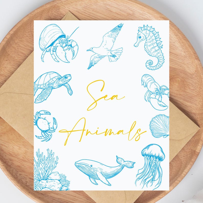 K&ouml;nnte beinhalten: Eine wei&szlig;e Karte mit blauen Illustrationen von Meerestieren, darunter eine Krabbe, eine Schildkr&ouml;te und ein Wal. Die Worte "Sea Animals" sind in gelber Schrift geschrieben. Die Karte liegt auf einem Holztablett mit einem braunen Umschlag.