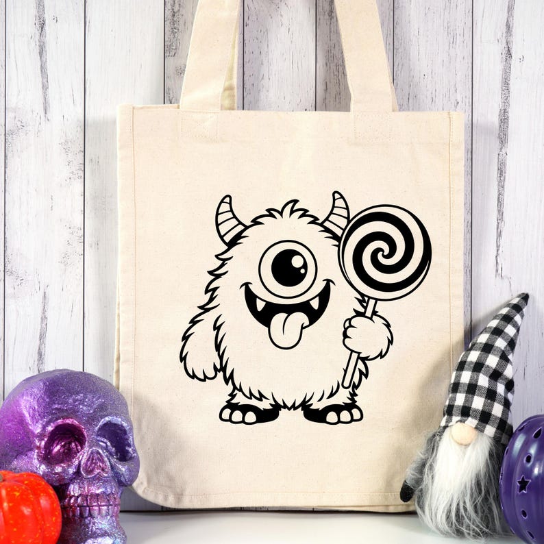 May include: A beige canvas tote bag featuring a black graphic print of a furry one-eyed monster with two horns, a wide smiling mouth with visible teeth, and a tongue sticking out while holding a large swirled lollipop in its hand.