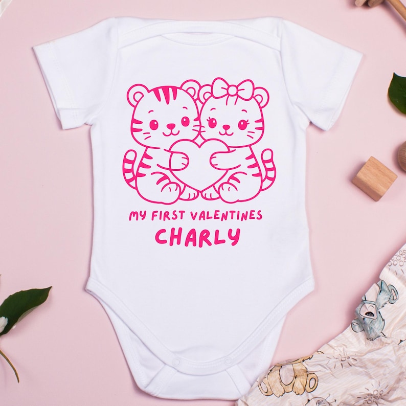 Puede incluir: Body de beb&eacute; blanco con un dise&ntilde;o rosa de dos cachorros de tigre abrazando un coraz&oacute;n. El texto "MY FIRST VALENTINES CHARLY" est&aacute; impreso debajo. La prenda es de material suave.