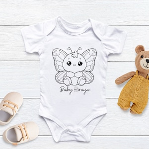 K&ouml;nnte beinhalten: Wei&szlig;er Baby-Body mit einem schwarz-wei&szlig;en Schmetterlingsmotiv und dem Text "Baby Hiraya". Ein Paar beige Babyschuhe und ein Stoffb&auml;r liegen neben dem Body.