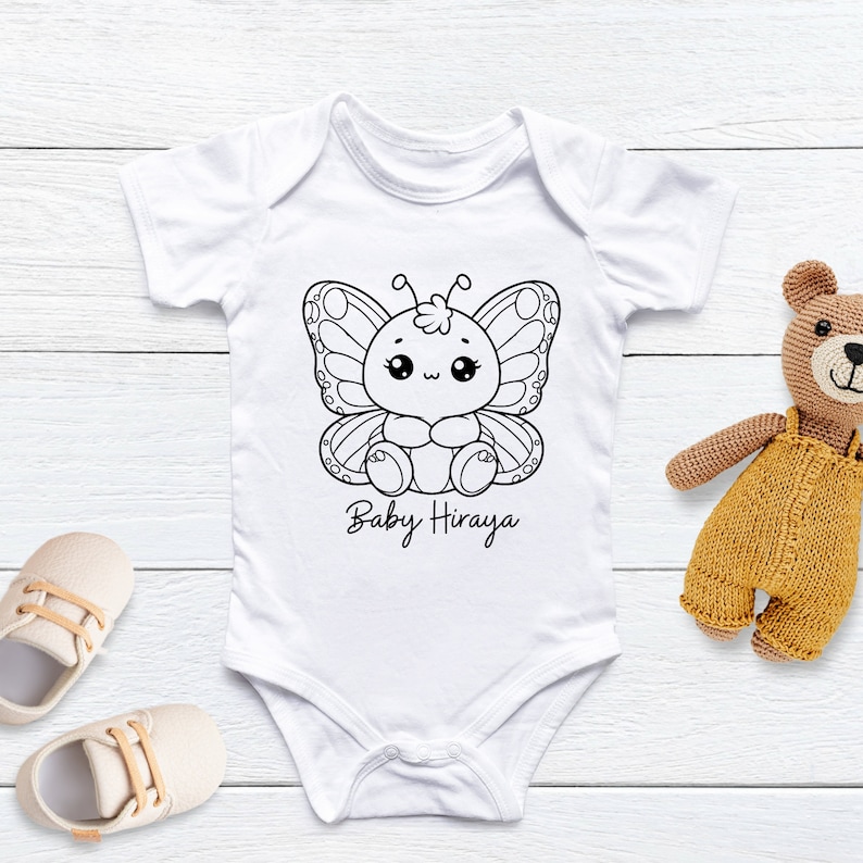 K&ouml;nnte beinhalten: Wei&szlig;er Baby-Body mit einem schwarz-wei&szlig;en Schmetterlingsmotiv und dem Text "Baby Hiraya". Ein Paar beige Babyschuhe und ein Stoffb&auml;r liegen neben dem Body.