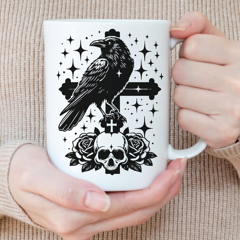 Pu&ograve; includere: Tazza in ceramica bianca con un disegno di corvo nero appollaiato su una croce sopra un teschio e rose, con accenti di stelle. La tazza ha un manico bianco. La tazza &egrave; tenuta da una persona.