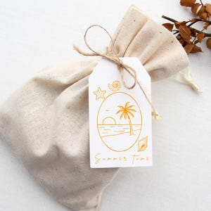 Pu&ograve; includere: Un sacchetto regalo in tessuto beige legato con spago, con un'etichetta bianca con una scena di spiaggia dorata. L'etichetta presenta una palma, il sole e le parole "Summer Time". Sono presenti anche conchiglie e una stella marina.